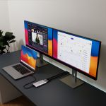 mbeat USB-C Dual 4K HDMI DisplayLink adapter connected to a MacBook Pro on a dark desk, driving two extended external monitors displaying a video call and an HR analytics dashboard in a modern home office setup.