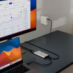 mbeat USB-C Dual 4K HDMI DisplayLink adapter on a dark desk, connected to a MacBook Pro and a large external monitor showing an analytics dashboard, with USB-C power pass-through cable running to a wall charger.